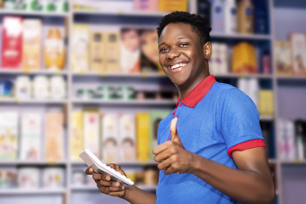 A smiling storekeeper at Karen Lux Wholesalers in Nairobi holding a calculator and giving a thumbs up inside a wholesale FMCG shop.